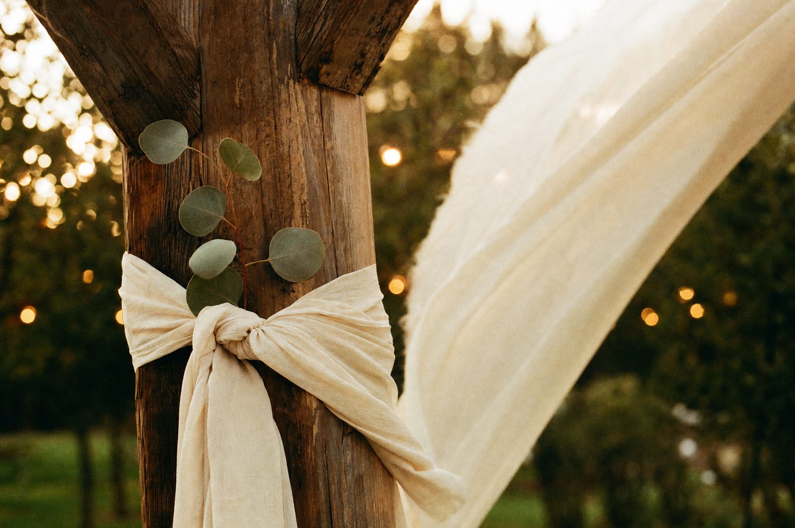 Ivory chiffon knotted twice around a rough wooden arch post with a sprig of eucalyptus tucked into the knot