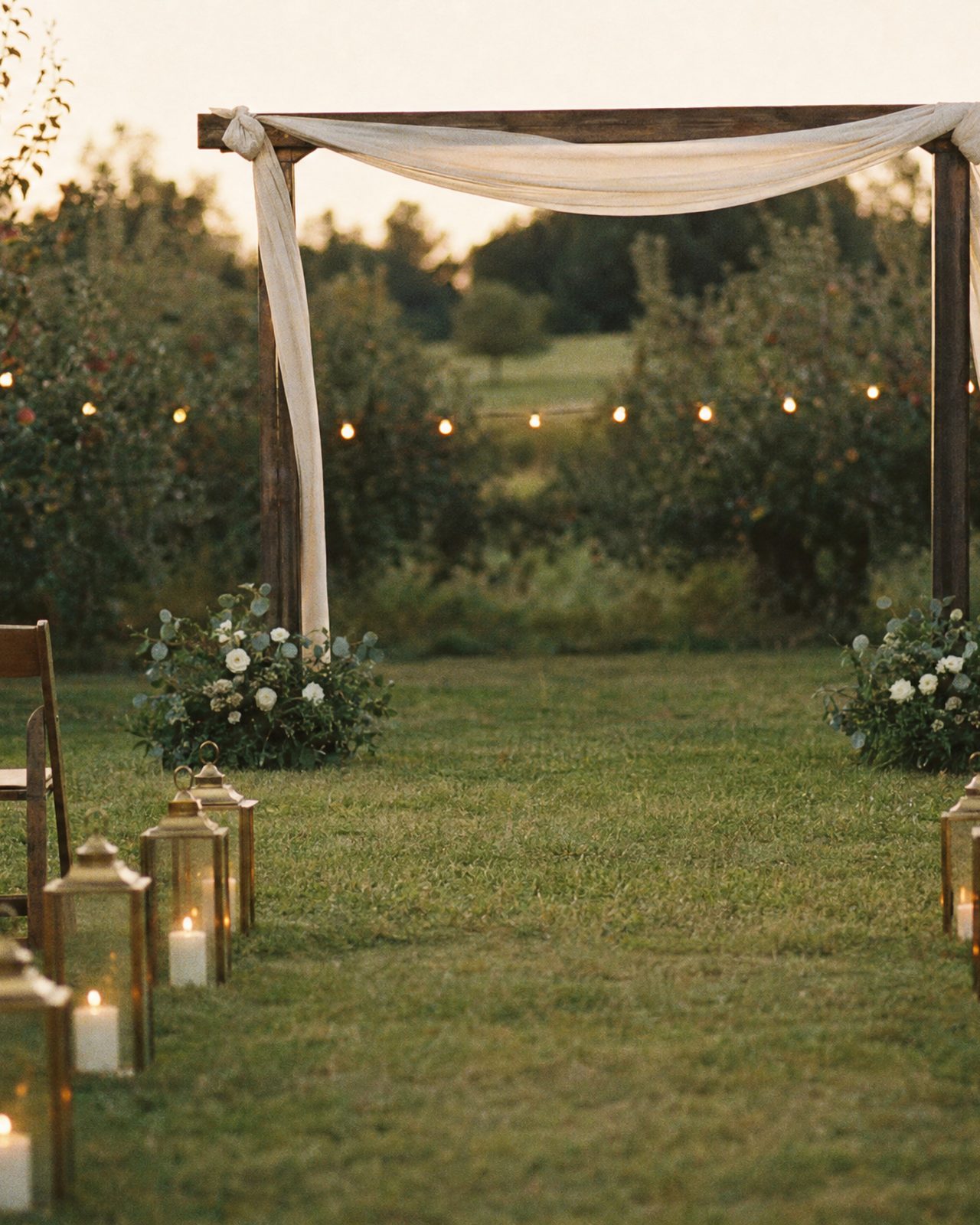 The orchard arch at dusk, chiffon moving in the wind, lanterns down the aisle