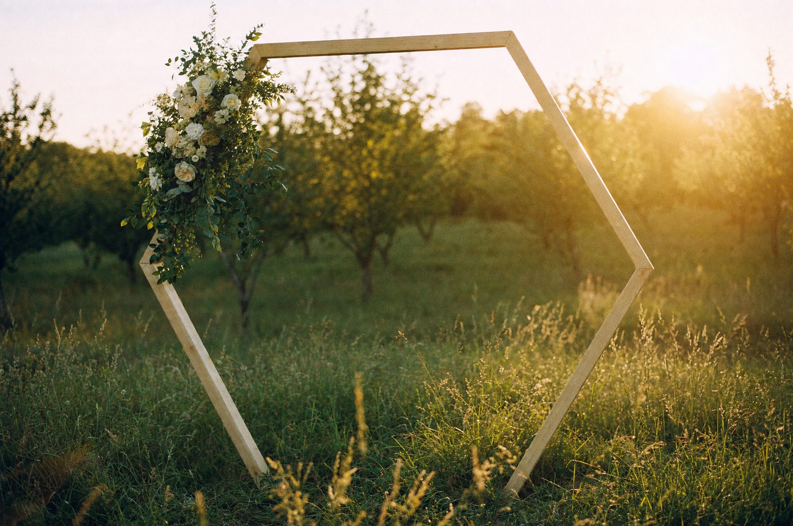 A bare wooden hexagon wedding arch in a farm meadow at golden hour with flowers on one upper corner only